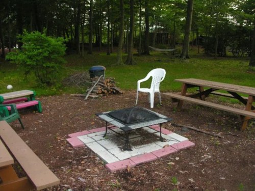A campsite with a fire pit, a white chair, picnic tables, and a stack of firewood surrounded by trees.