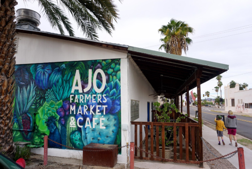 Colorful mural of "Ajo Farmers Market & Cafe" on a building, with palm trees and pedestrians nearby.