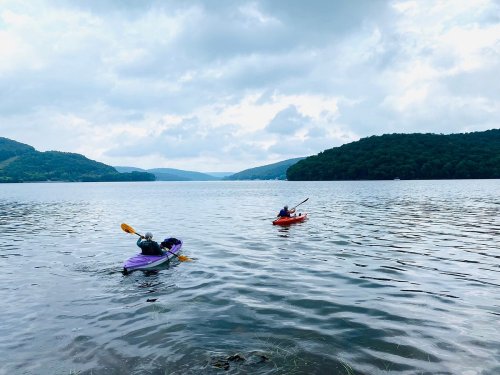 Two kayakers paddle on a calm lake surrounded by green hills under a cloudy sky.