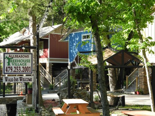 A sign for Allseasons Treehouse Village with colorful treehouses and a picnic area surrounded by greenery.