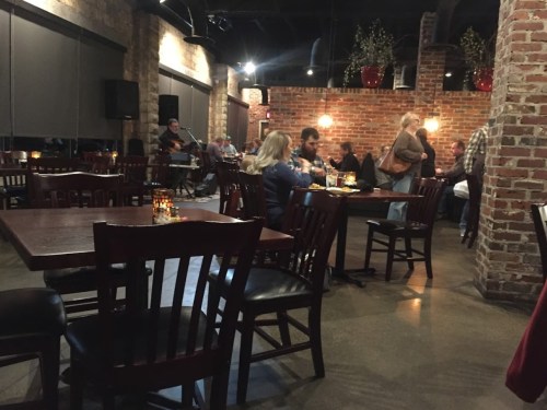 A cozy restaurant interior with wooden tables, brick walls, and a small live band performing in the background.