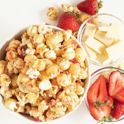 A bowl of popcorn mixed with strawberries, alongside bowls of white chocolate and fresh strawberries.