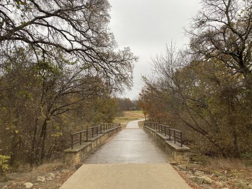 A stone bridge leads to a path through trees on a cloudy day, with autumn foliage visible along the sides.