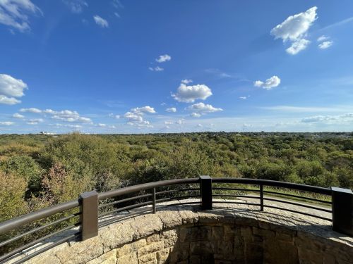 A scenic view of lush green trees under a blue sky with fluffy clouds, seen from a stone railing.