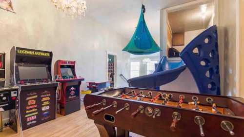 A game room featuring a foosball table, arcade machines, and a slide, with bright decor and a chandelier.