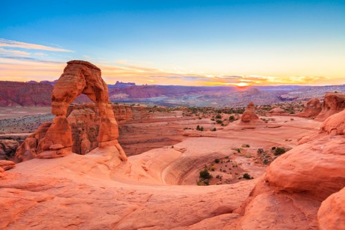 Sunset over Arches National Park, featuring the iconic Delicate Arch amidst red rock formations and a colorful sky.