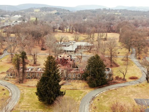 Aerial view of an abandoned building surrounded by trees and open fields, with mountains in the background.