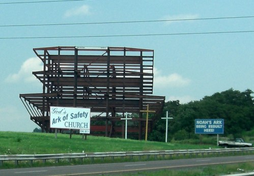 A partially constructed steel structure with signs for "God's Ark of Safety Church" and "Noah's Ark being rebuilt here."