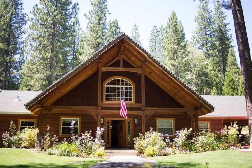 A rustic wooden house with a triangular roof, surrounded by tall trees and colorful flowers, featuring an American flag.