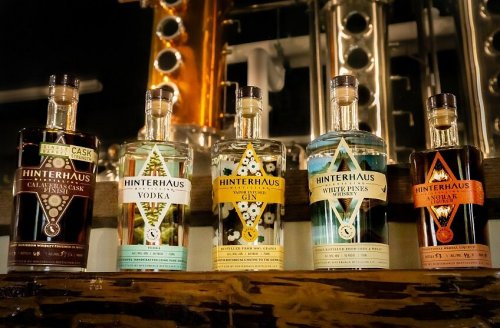 Five bottles of Hinterhaus spirits lined up on a wooden shelf, showcasing various flavors and designs against a distillery backdrop.