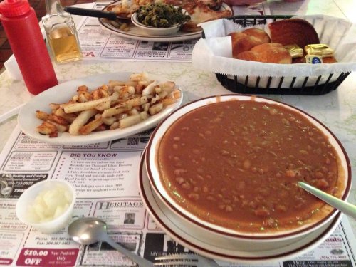 A bowl of brown beans with a spoon, served with fried potatoes, cornbread, and condiments on a restaurant table.