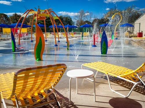 Colorful splash pad with water features, yellow lounge chairs, and blue umbrellas under a clear sky.
