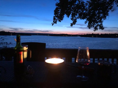 A serene lakeside view at dusk, featuring a candle, wine glass, and a bottle, with a colorful sky reflecting on the water.