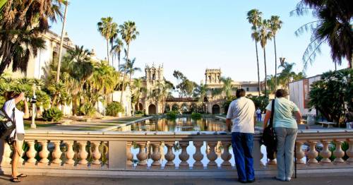 Two people admire a scenic view of a palm-lined garden and pond, with historic buildings in the background.