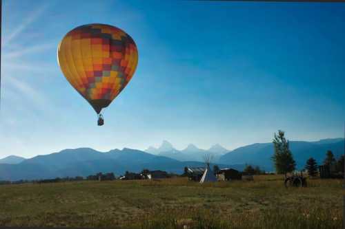 A colorful hot air balloon floats over a scenic landscape with mountains and a teepee under a clear blue sky.