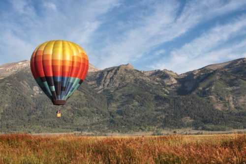 A colorful hot air balloon floats over a grassy field with mountains in the background under a blue sky.