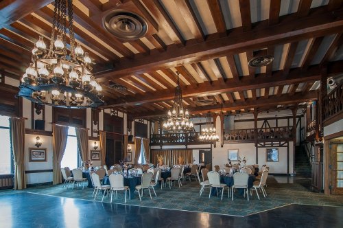 Elegant banquet hall with wooden beams, chandeliers, and round tables set for dining. Warm, inviting atmosphere.
