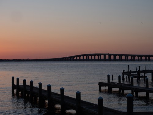 A serene sunset over a calm body of water, featuring a bridge and wooden piers in silhouette.