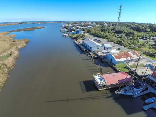 Aerial view of a calm river with boats, docks, and buildings along the shore under a clear blue sky.