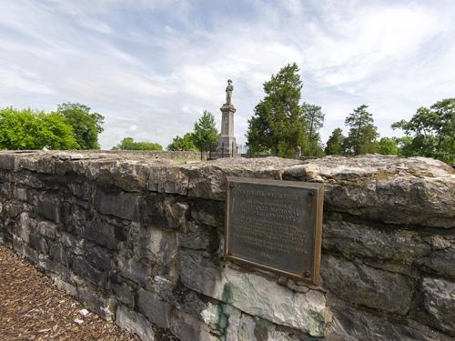 A stone wall with a plaque in front of a tall monument surrounded by trees under a cloudy sky.