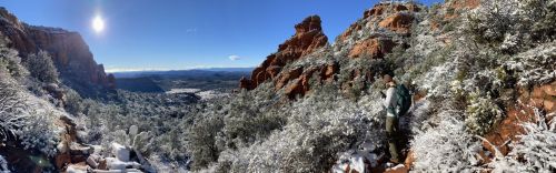 A hiker stands on a snowy trail surrounded by red rock formations and evergreen trees under a clear blue sky.