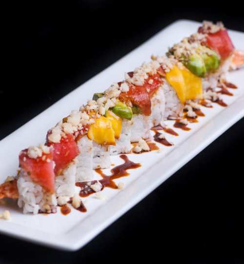 A colorful sushi roll topped with mango, tuna, and avocado, garnished with crushed nuts and drizzled with sauce.