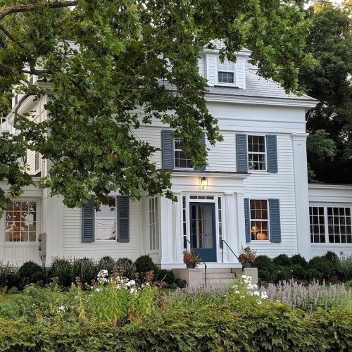 A charming white house with blue shutters, surrounded by greenery and colorful flowers in a peaceful setting.