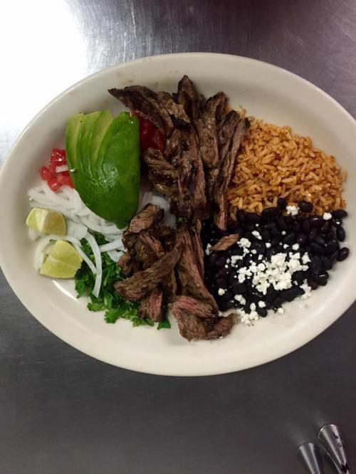A plate of grilled steak with rice, black beans, avocado, tomatoes, onions, cilantro, and lime wedges.