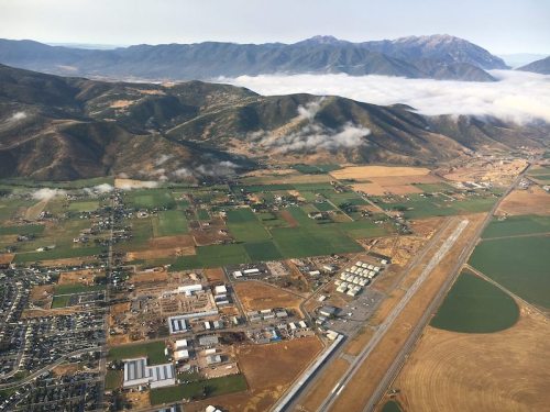 Aerial view of a rural landscape with fields, buildings, and mountains in the background, partially covered by clouds.