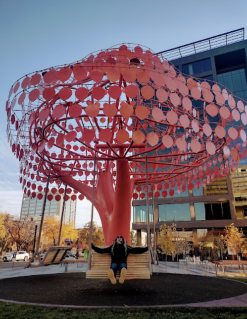 A person swings beneath a large, vibrant red tree sculpture with circular leaves against a clear blue sky.