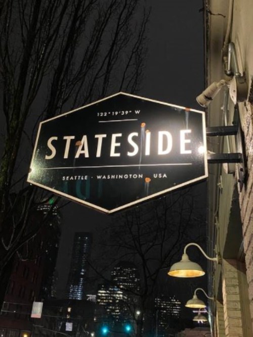 Sign for "Stateside" restaurant in Seattle, Washington, illuminated at night with city buildings in the background.