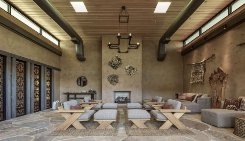 A spacious, modern living room with stone flooring, wooden beams, and cozy seating around a central fireplace.