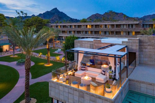 A luxurious outdoor lounge area with fire pits, surrounded by palm trees and mountains at dusk.
