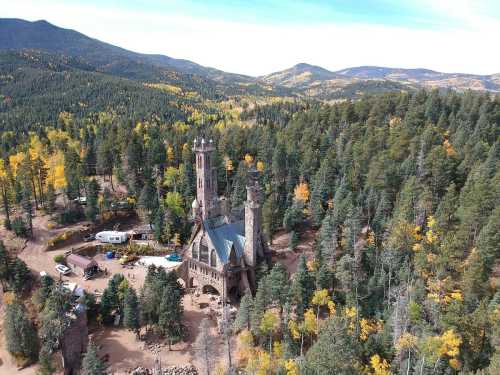 Aerial view of a castle-like structure surrounded by dense forests and mountains, with autumn foliage in the landscape.