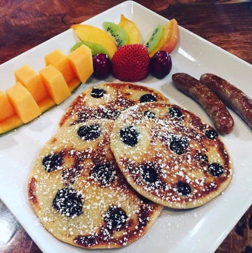 Plate of blueberry pancakes dusted with powdered sugar, served with fresh fruit and sausage links.