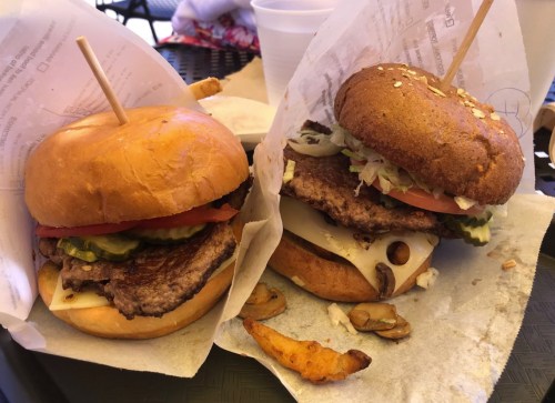 Two gourmet burgers on paper wrappers, one with lettuce, tomato, pickles, and cheese, alongside a few fries.