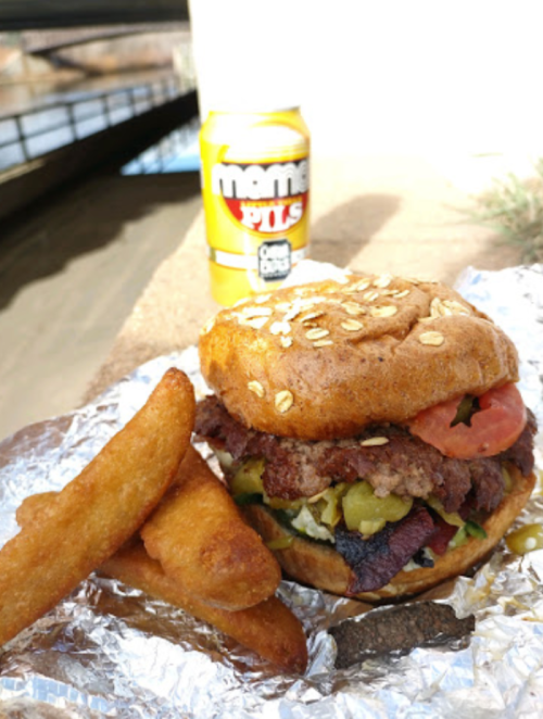 A close-up of a burger with lettuce, tomato, and pickles, accompanied by fried snacks and a can of beer.
