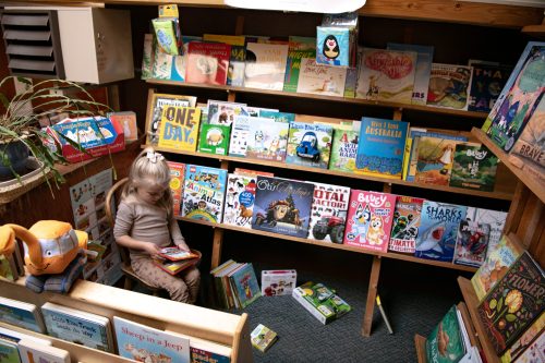 A child sits on a bench in a cozy book nook, surrounded by colorful children's books on shelves.