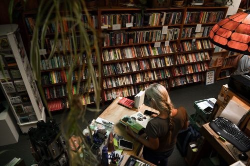 A person browses books at a cozy bookstore filled with shelves of colorful books and a warm lamp.
