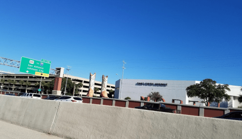 View of a highway sign directing to San Antonio, with a shopping center and parking garage in the background under a clear blue sky.
