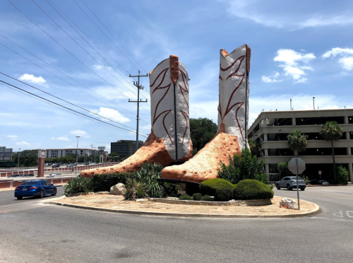 Giant cowboy boots sculpture in a roundabout, surrounded by greenery and a blue sky.