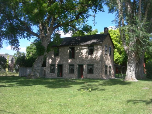 A stone house surrounded by large trees and green grass under a clear blue sky.