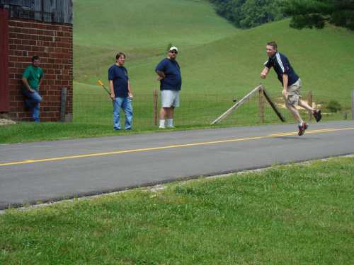 A man runs on a road while three others watch from the side, with green hills in the background.