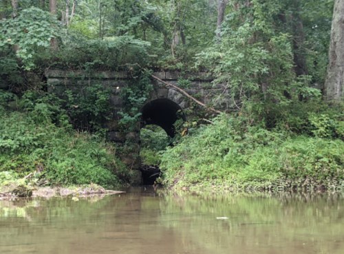 A stone arch bridge over a calm stream, surrounded by lush greenery and trees.