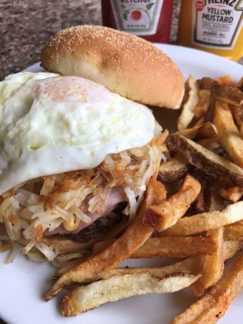 A plate with a burger topped with a fried egg and hash browns, served with a side of crispy French fries.
