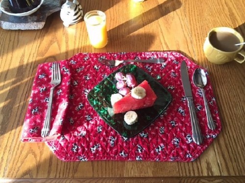 A colorful table setting with a red floral placemat, featuring a plate of fruit, utensils, and a drink.
