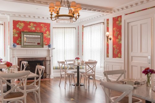 A cozy dining room with floral wallpaper, a chandelier, and tables set for guests near a fireplace.