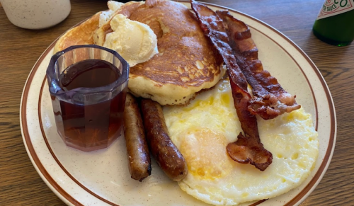 A plate of breakfast featuring pancakes, eggs, bacon, sausage, and a glass of syrup.