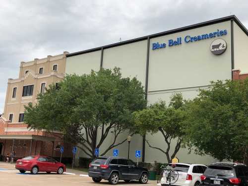 Exterior of Blue Bell Creameries building with trees and parked cars in front, under a cloudy sky.