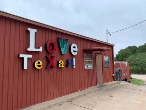 Colorful sign reading "Love Texas!" on a red metal building, with a cloudy sky in the background.